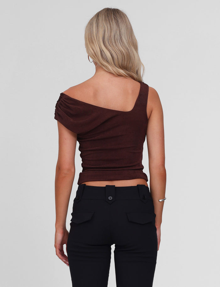 A woman with blonde wavy hair faces away, wearing the TIGERMIST CASSIAN TOP in chocolate and high-waisted black pants against a plain light background.