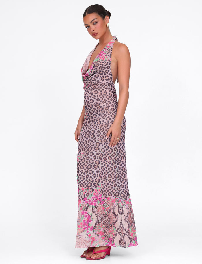 A woman models the TIGERMIST CATALINA MAXI DRESS - NEUTRAL : ANIMAL : ANIMAL FLORAL BORDER, a sleeveless, backless animal print dress with a floral border, paired with pink heels against a plain white background.