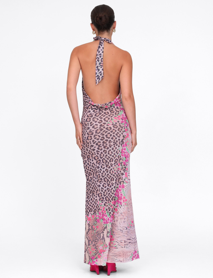 A woman with her hair in a bun models the TIGERMIST CATALINA MAXI DRESS in Neutral Animal Floral Border, showcasing its backless design and pink shoes while facing away against a plain white background.