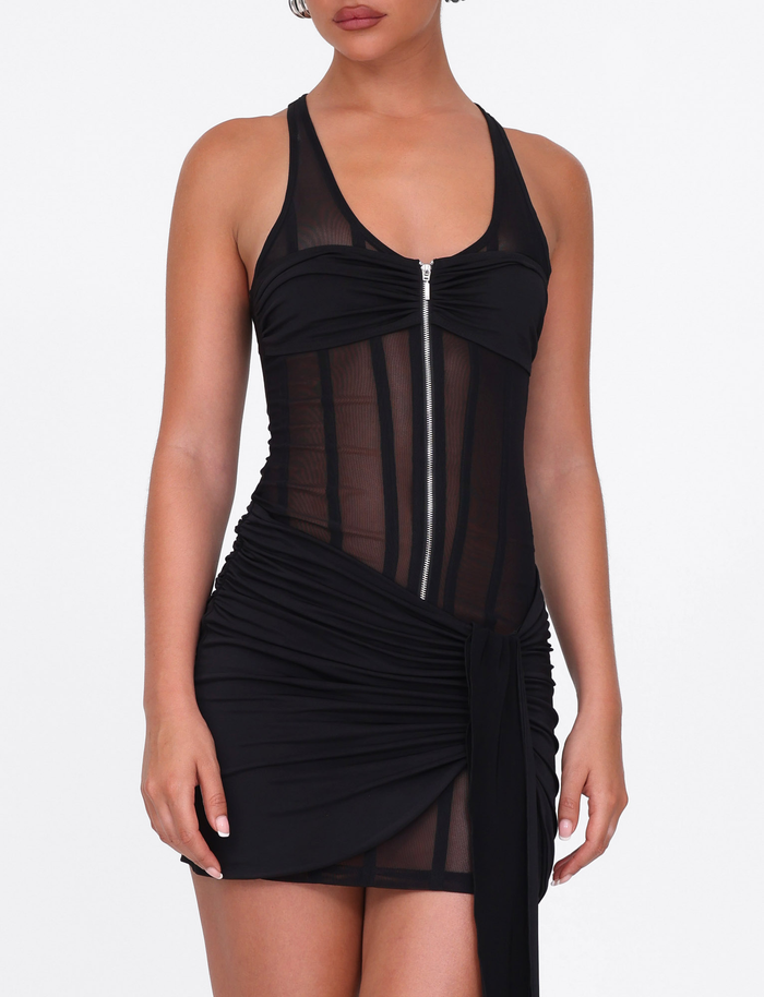 A person wears the TIGERMIST CATERINA MINI DRESS in black, featuring a fitted mesh design with sheer panel lines, front zipper, and an asymmetrical draped skirt, standing against a plain white background.