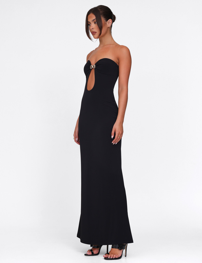 A woman models the TIGERMIST CECILY MAXI DRESS - BLACK, a strapless, floor-length gown with a sweetheart neckline, front cutout, and circular bust detail. She pairs it with high-heeled sandals against a plain white background.