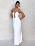A woman poses against a light textured wall, wearing the TIGERMIST CECILY MAXI DRESS in white—a strapless style featuring a front keyhole cutout with a gold ring. Her dark hair is styled in a bun with face-framing strands, hands behind her back.