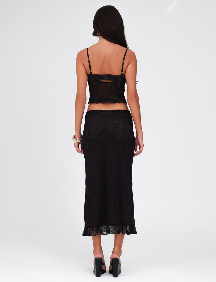 A woman faces away from the camera in a TIGERMIST CHARLEIGH SKIRT - BLACK : LACE, paired with a black lace camisole, high-heeled shoes, and a bracelet against a plain white background.