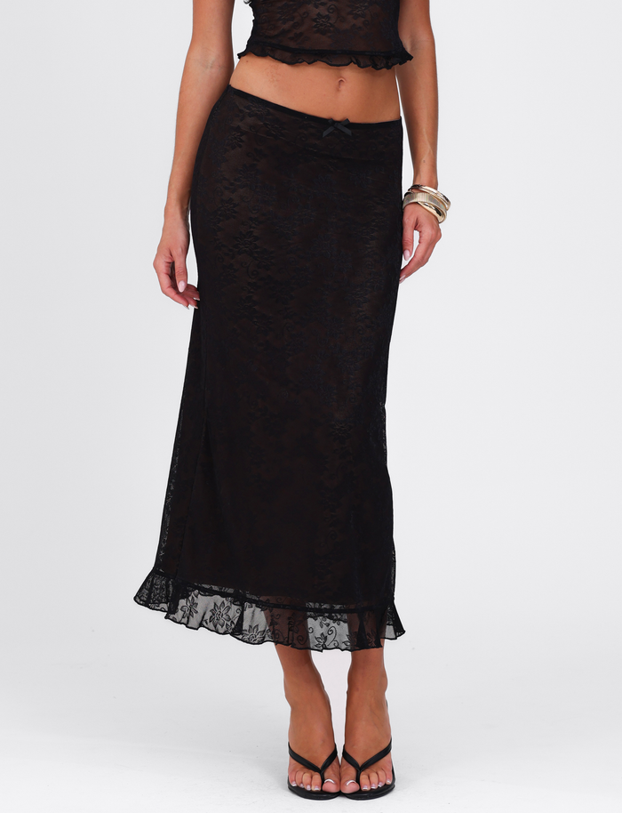 A woman wearing the TIGERMIST CHARLEIGH SKIRT in black lace with a ruffled hem, paired with a matching sleeveless top, black sandals, and gold bangles stands against a plain white background.