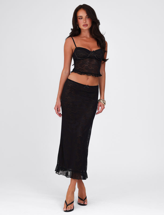 A woman with long, wavy dark hair wears the TIGERMIST CHARLEIGH SKIRT - BLACK : LACE in a midi style, paired with a sheer black lace tank top, black heeled sandals, and gold bracelets against a plain white background.