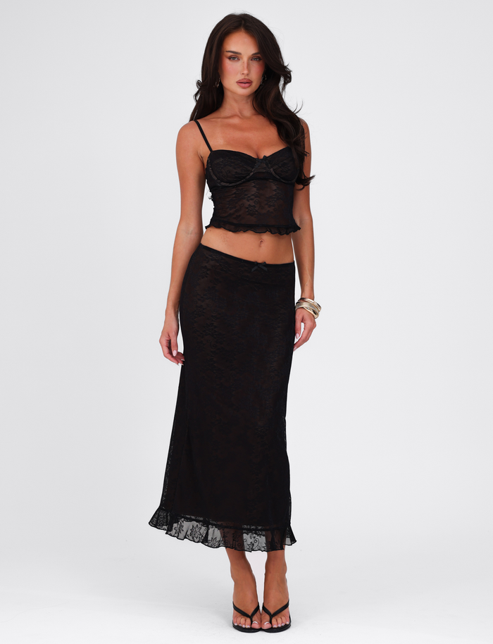 A woman stands against a white background wearing the TIGERMIST CHARLEIGH TOP in black lace and a matching skirt, styled with black sandals and gold bracelets on her right wrist.