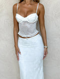A woman stands against a beige textured wall wearing the CL - CHARLEIGH TOP by Tiger Mist USA—a fitted, white, floral lace camisole—paired with a matching high-waisted maxi skirt and bracelets on her left wrist.