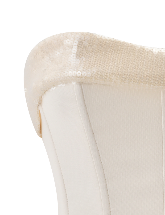 Close-up of a white upholstered chair's backrest adorned with the TIGERMIST CHARLYZE BUSTIER in cream, its sequined details draped elegantly over the top edge, evoking the refined style of a corset bustier.