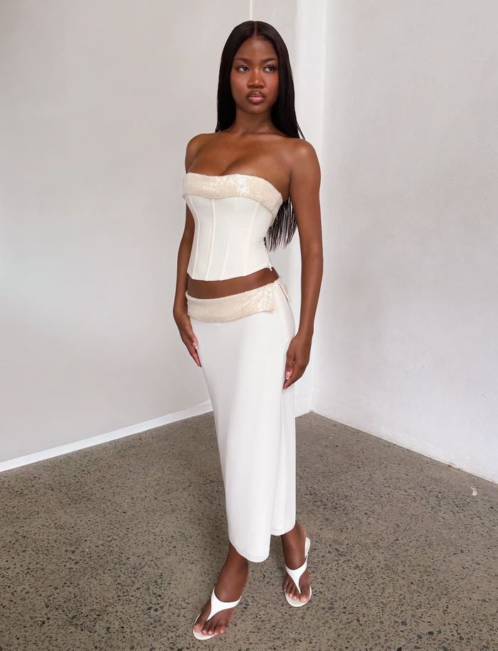 A woman stands indoors wearing the TIGERMIST CHARLYZE MIDI SKIRT in cream, paired with a strapless corset top with gold accents, white heeled sandals, and has long straight hair. The setting features white walls and a concrete floor.