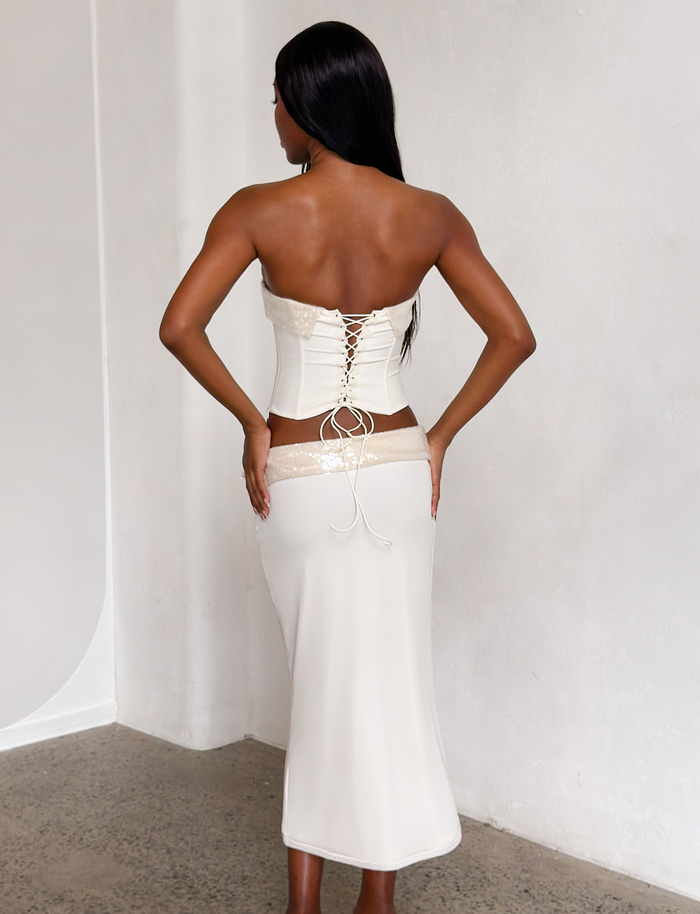 A woman stands with her back to the camera in a minimalist, light-filled room, wearing the TIGERMIST CHARLYZE MIDI SKIRT in cream and a strapless white lace-up corset, highlighting her figure against the concrete floor and white walls.