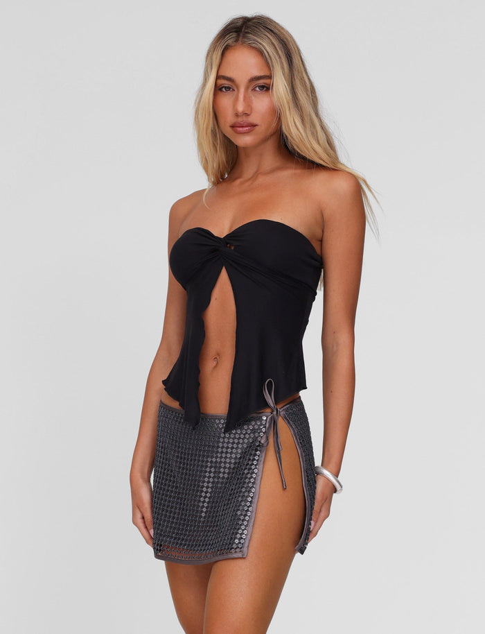 A woman with long blonde hair models the TIGERMIST CHARMAINE TOP in black, a strapless front-tie design, paired with a short textured gray skirt featuring a twist detail, standing against a plain white background.