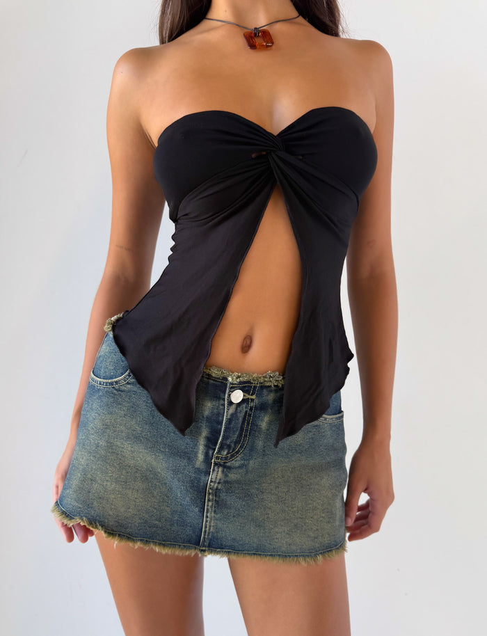 A woman stands against a plain light background wearing the TIGERMIST CHARMAINE TOP in black, a strapless design featuring a front cutout and knot detail, paired with a frayed denim mini skirt.