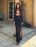 A woman stands on a stone patio in front of a glass door, wearing the CHRISTY LACE L/S MAXI DRESS - BLACK : LACE by TIGERMIST, which features a cutout at the front and a high slit. She has long, wavy hair and wears black open-toe heels. A stone bust and potted plant are in the background.