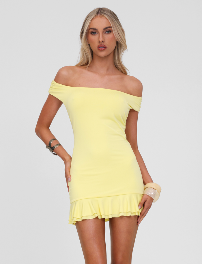 A woman poses against a white background wearing the TIGERMIST CLIA MINI DRESS in lemon, an off-shoulder mini dress with a frill hemline. She has long blonde hair and accessorizes with bracelets on both wrists.