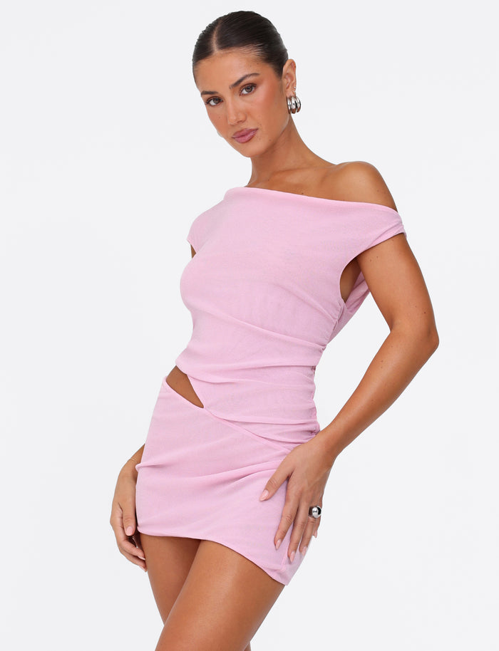 A woman poses confidently in the TIGERMIST COBI MINI DRESS 2.0 - BABY PINK, featuring an off-the-shoulder, sleeveless design with a side cut-out. Her hair is pulled back, styled with hoop earrings and a ring against a plain white background.