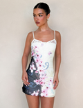 A woman stands against a plain background in the TIGERMIST COLSON MESH MINI DRESS - MULTI: FLORAL: CHERRY BLOSSOM, featuring adjustable straps. She has slicked-back hair, pearl earrings, and looks to the side.