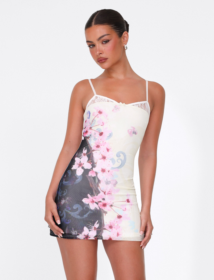 A woman with dark hair in a sleek bun models the TIGERMIST COLSON MESH MINI DRESS – MULTI: FLORAL: CHERRY BLOSSOM, featuring adjustable straps and a cream, black, and pink cherry blossom floral design, against a plain white background.
