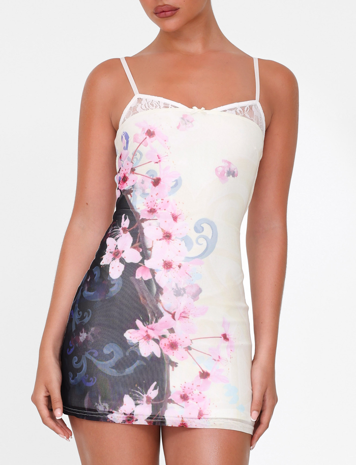 A woman wears the TIGERMIST COLSON MESH MINI DRESS - MULTI, featuring a body-hugging fit, adjustable straps, lace-trimmed neckline, and cherry blossom floral print on a white and black gradient background.
