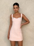 A woman in the TIGERMIST COLSON MINI DRESS in baby pink, featuring wide straps and white lace trim, poses against a textured beige wall with her hand on her hip, looking at the camera.
