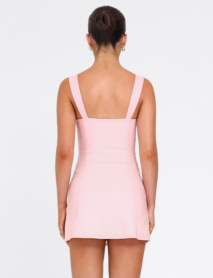 A woman with dark hair in a neat bun is seen from behind, wearing the TIGERMIST COLSON MINI DRESS in baby pink, a sleeveless fitted mini dress with wide shoulder straps, against a plain white background.