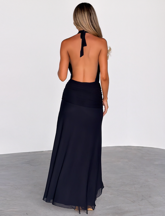 A woman with long, wavy hair stands with her back to the camera, wearing the TIGERMIST CORELLE DRESS in black—a floor-length plunge maxi—on a plain gray floor against a white wall background.