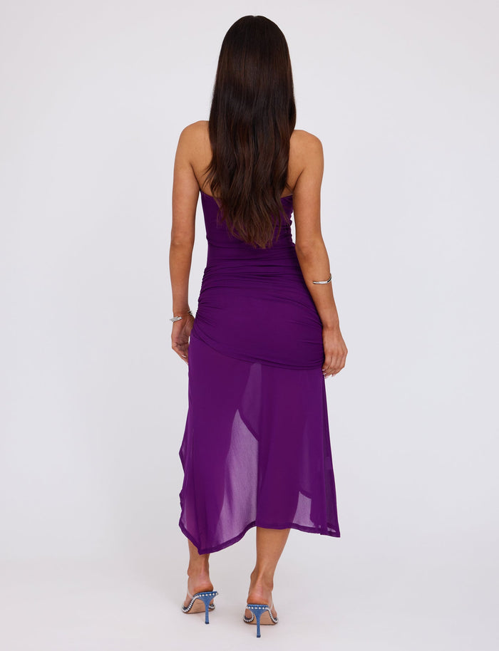 A woman stands with her back to the camera, wearing the COVE DRESS in purple from TIGERMIST. The strapless dress is fitted at the top and flows down to a mid-calf length. She has long, dark hair and is wearing strappy blue high-heeled sandals. The background is plain white.
