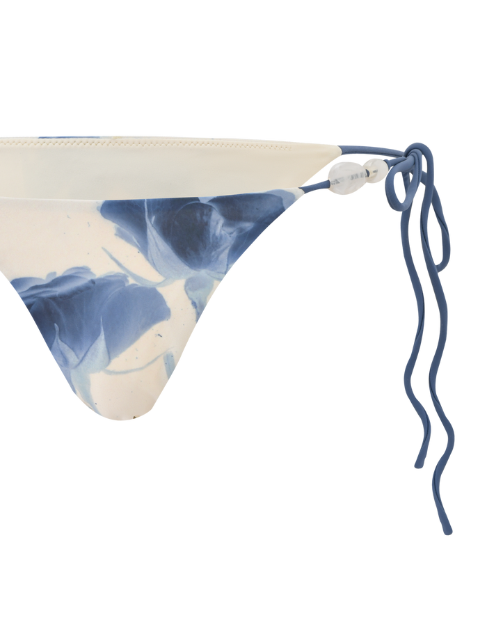 The CYLAS BIKINI BOTTOM - INDIGO FLORAL by TIGERMIST features a blue and white floral pattern, displayed from a side angle on a white background.
