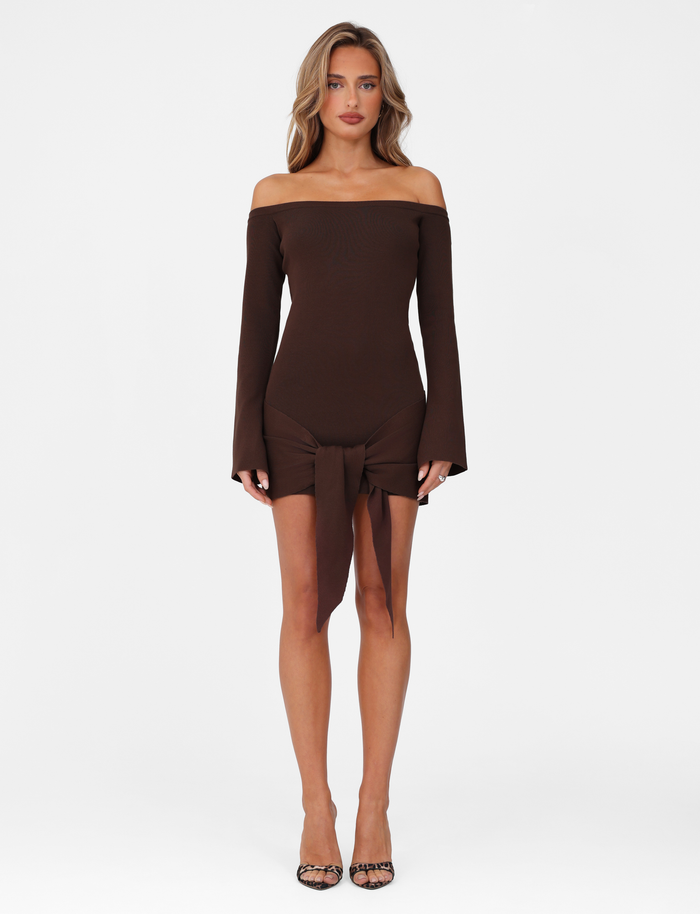A woman with wavy hair and a neutral expression stands against a white background, wearing the TIGERMIST CYPRESS MINI DRESS in chocolate brown. The dress features an off-the-shoulder neckline, long sleeves, and a front tie detail, paired with open-toe heels.
