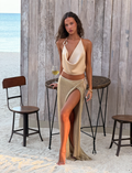 A woman in the TIGERMIST CYRENE TIE MAXI SKIRT - GOLD stands barefoot on sand, holding a wine glass by a wooden table and chairs, with a wooden backdrop at the beach.