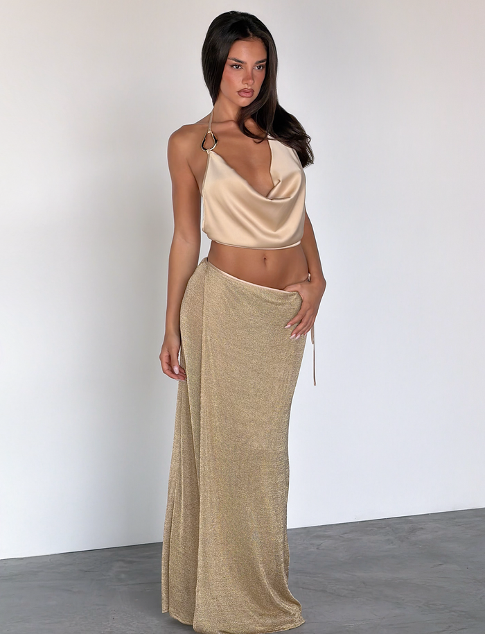 A woman with long, dark hair poses indoors against a white wall, wearing the TIGERMIST CYRENE TIE MAXI SKIRT in gold and a matching shimmery gold halter top, with one hand on her hip.