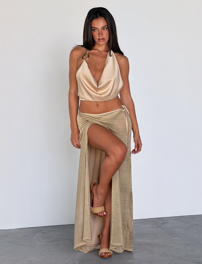 A woman with straight dark hair and a confident expression stands against a plain wall, wearing the TIGERMIST CYRENE TIE MAXI SKIRT in gold, paired with a shimmery halter top and gold high-heeled sandals.