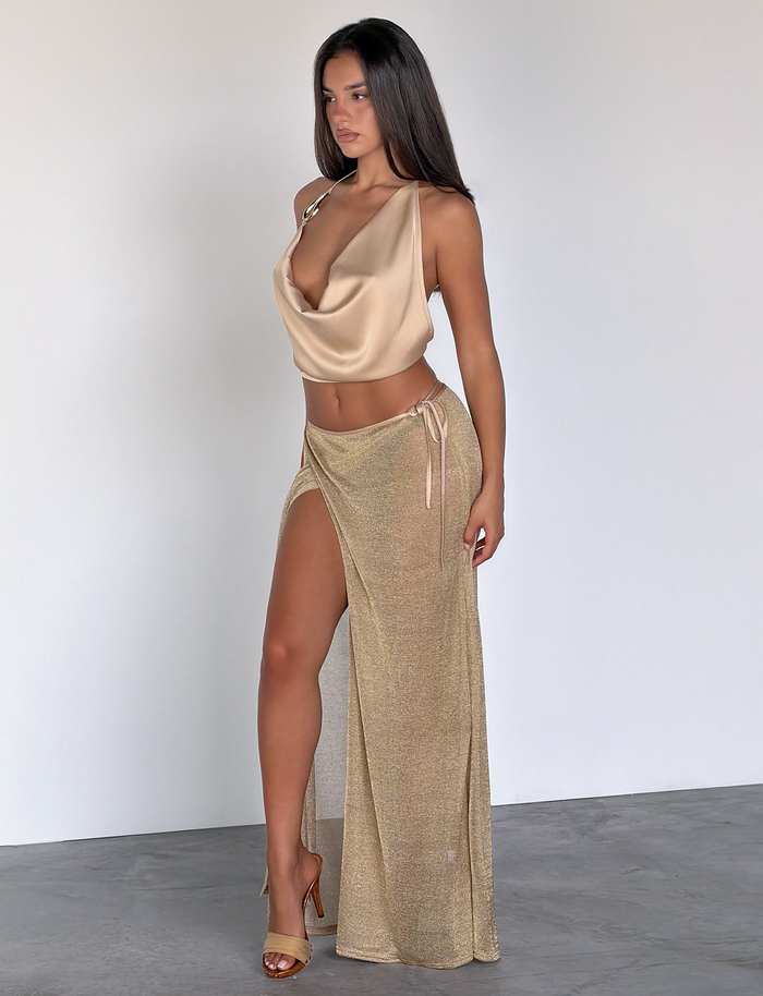 A woman stands indoors against a light wall, wearing the TIGERMIST CYRENE TIE MAXI SKIRT in gold paired with matching heels and a silky champagne draped top with satin trim.