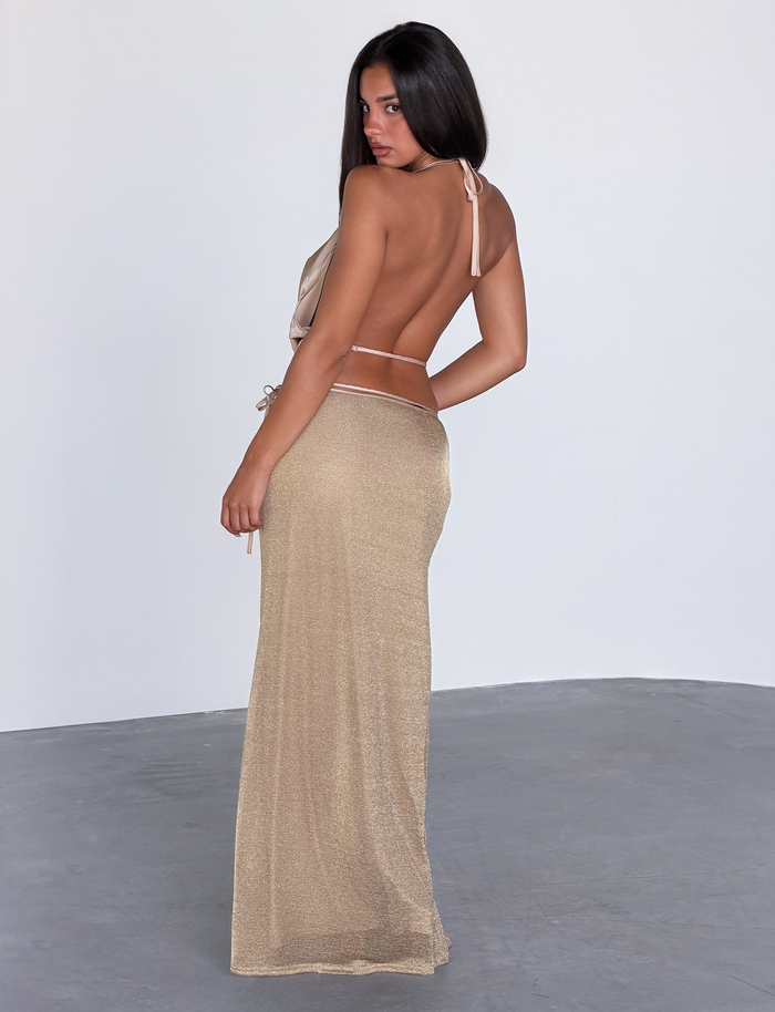 A woman with long dark hair stands in a minimalist room, wearing the TIGERMIST CYRENE TIE MAXI SKIRT in gold, featuring thin straps and a low-cut back. She looks over her shoulder toward the camera.