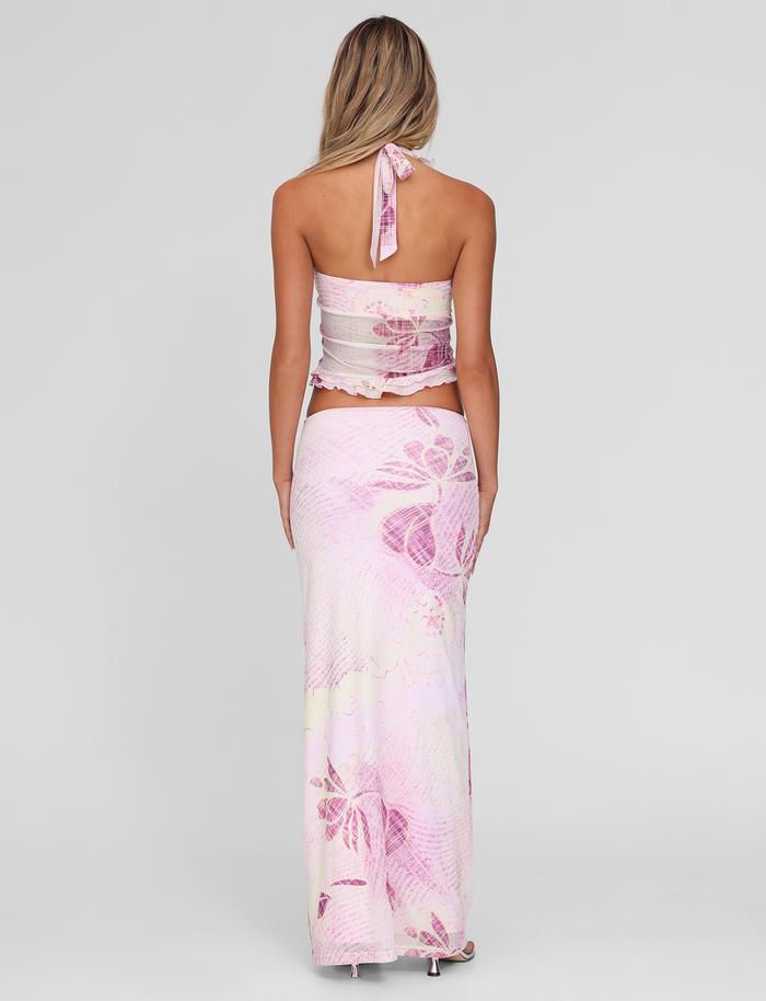 A woman with long blonde hair stands facing away, wearing the TIGERMIST DALILA MIDI SKIRT in Grunge Floral Magenta and a matching pink halter-neck crop top. The background is plain light gray.