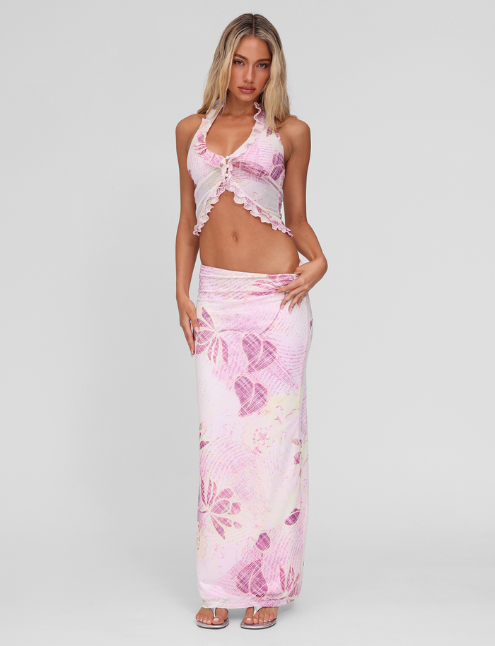A woman poses in the TIGERMIST DALILA TOP - GRUNGE FLORAL MAGENTA, a sleeveless ruffled halter crop top with a matching long skirt, featuring a pink and white floral pattern against a plain light gray background.