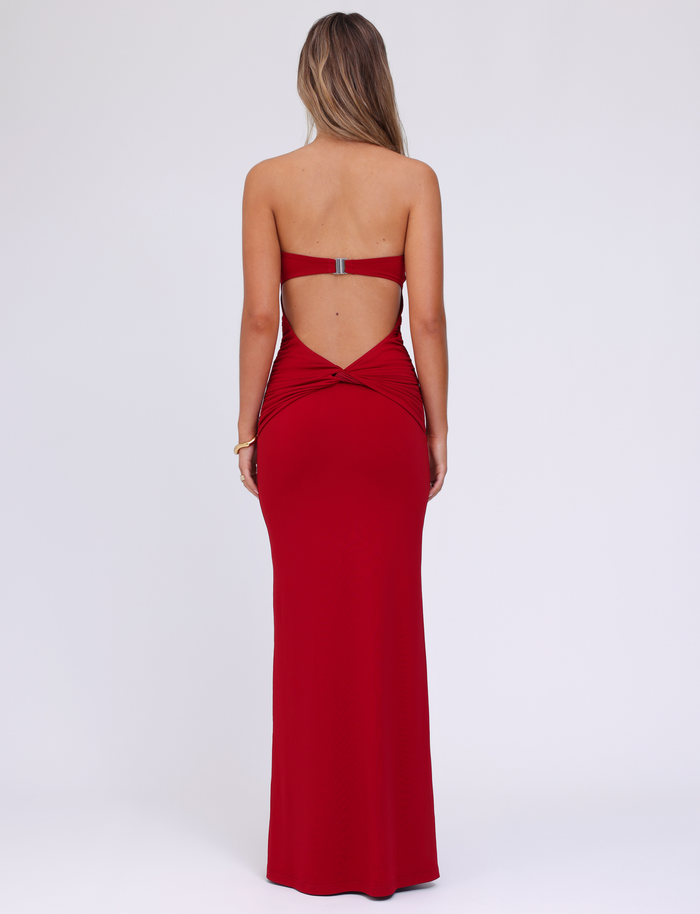 A woman with loose hair faces away in the TIGERMIST DANIELA DRESS - RED, a strapless soft jersey dress featuring an open back and upper back clasp, set against a plain white background.