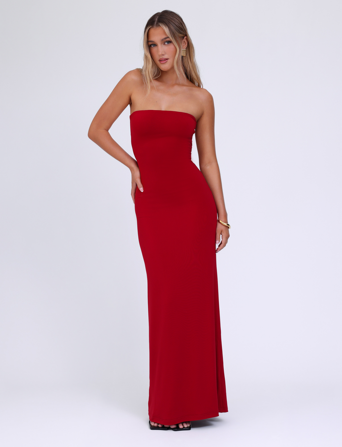 A woman wears the TIGERMIST DANIELA DRESS in red, a strapless, fitted maxi dress made from jersey. She stands against a plain white background, with long wavy blonde hair, black heels, and one hand on her hip.