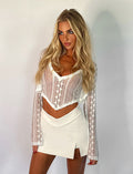 A blonde woman poses against a plain white background, wearing a sheer white lace long-sleeved top with a deep V-neck and the DAPHNIE PU SKORT - WHITE from TIGERMIST. Her hair is styled in loose waves, and she looks confidently at the camera.