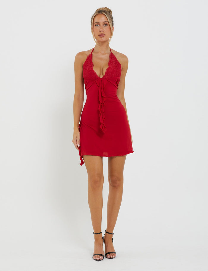 A woman stands against a white background wearing the DEENA DRESS - RED from TIGERMIST, a short red dress with a halter neckline and intricate lace details. The dress features a ruffle design down the front. She is wearing black high-heeled sandals and has her hair styled in an updo.