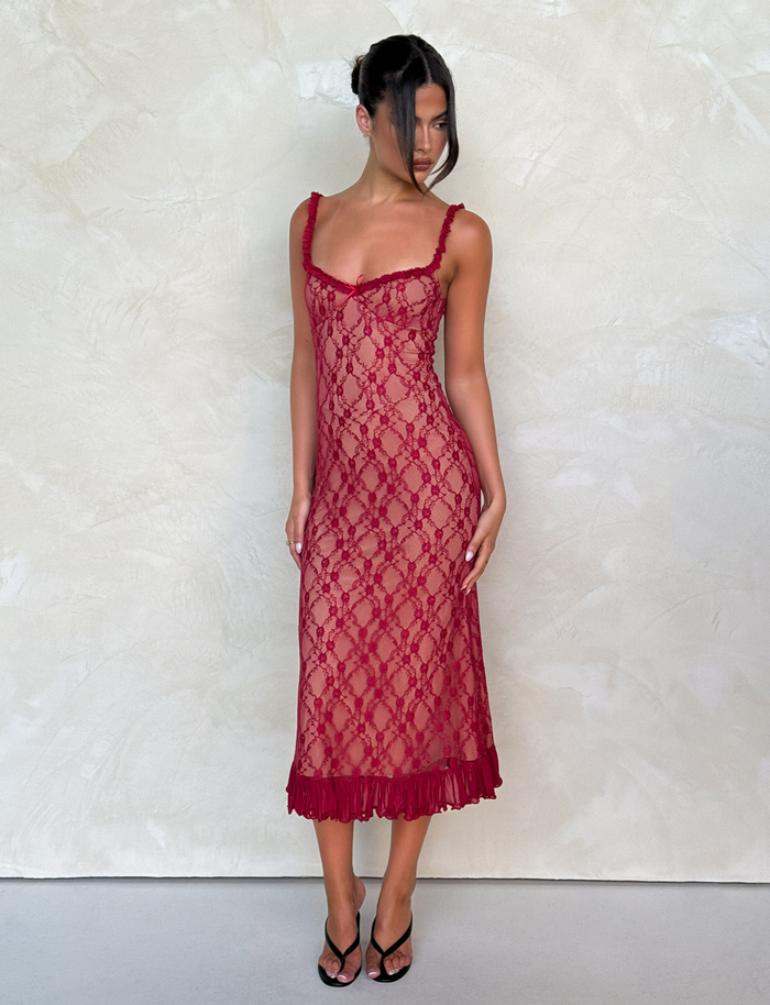 A woman in the TIGERMIST DEJA DRESS - RED : BERRY RED, a fitted sleeveless lace midi dress with a ruffled hem, stands against a light beige wall. She pairs it with black strappy heels and an elegant updo, gazing down and to the side.