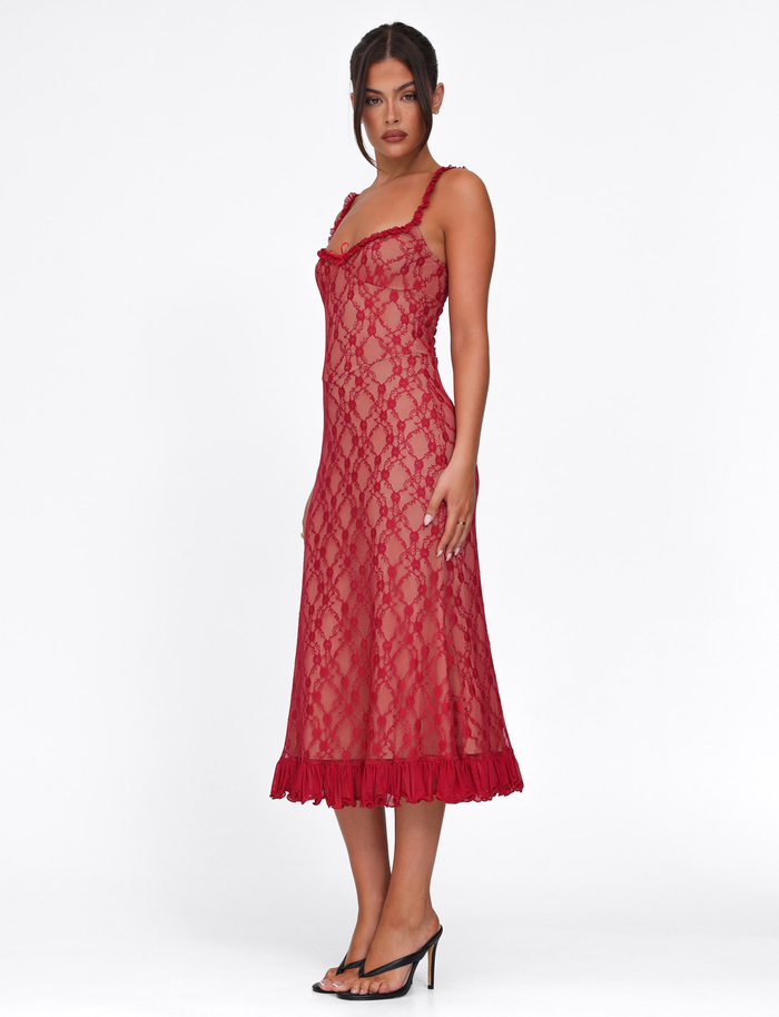 A woman with dark hair in a loose updo stands against a white background wearing the TIGERMIST DEJA DRESS - RED: BERRY RED, a semi-sheer lace midi dress with thin straps and a ruffled hem, paired with black heeled sandals.