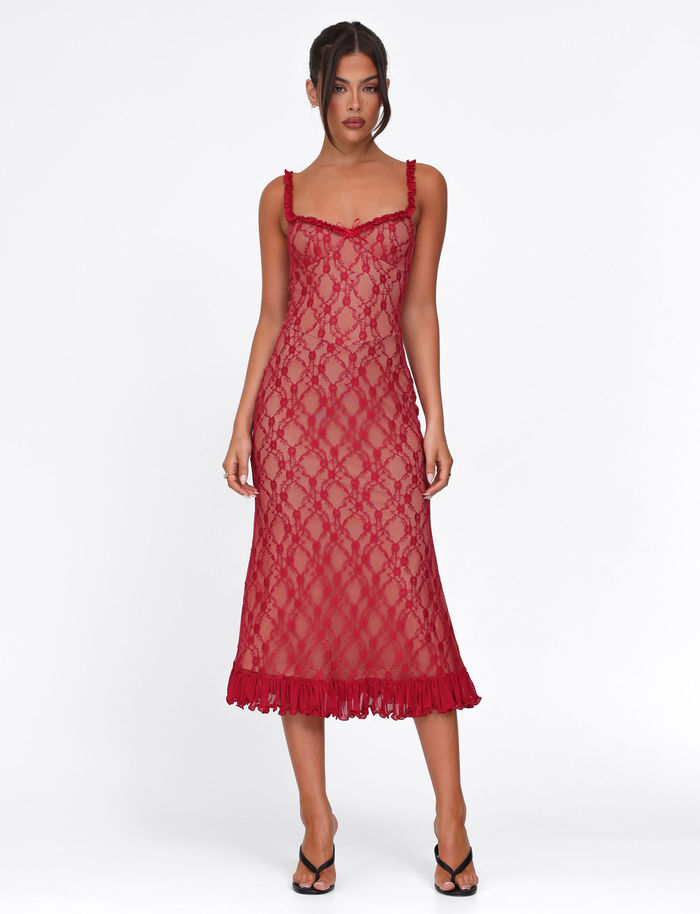 A woman stands against a white background wearing the TIGERMIST DEJA DRESS in Berry Red—a sleeveless, sheer midi dress with lace pattern and ruffled hem—paired with black heeled sandals.