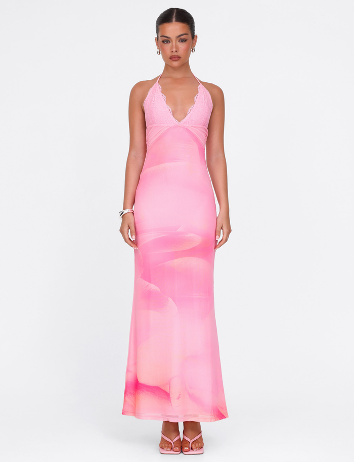 A woman wears the TIGERMIST DENAE MAXI DRESS in Pink Blossom, featuring a fitted silhouette, halter neckline, deep V cut, and subtle floral lace pattern. She styles it with pink sandals, simple jewelry, and a pulled-back hairstyle against a white background.