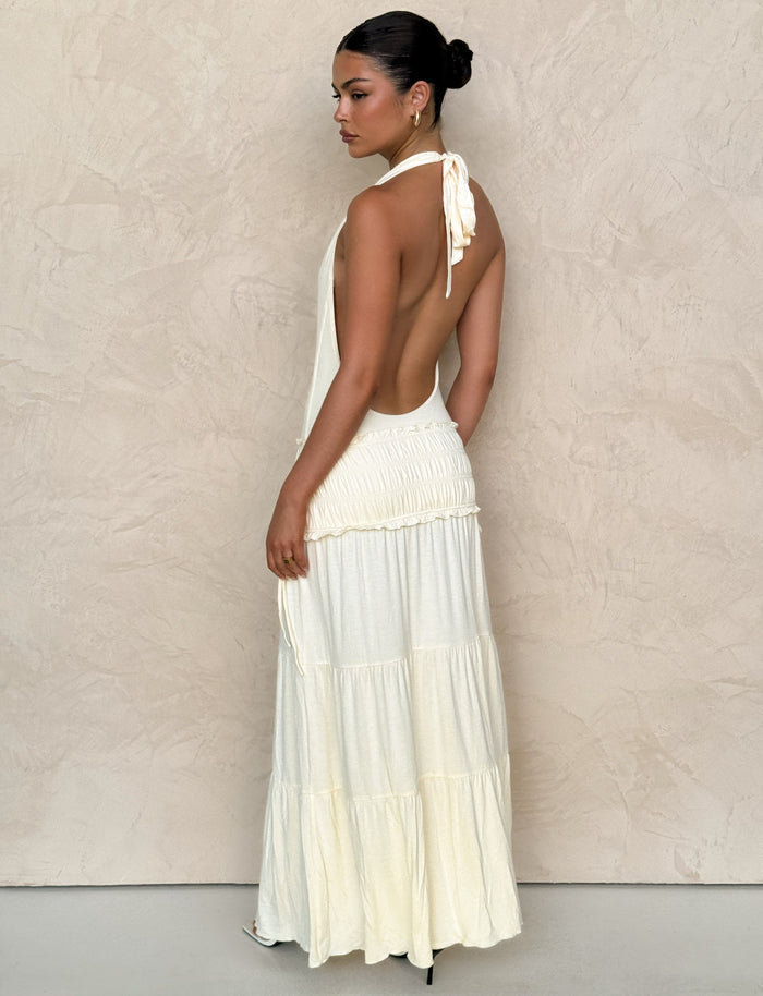 A woman poses sideways against a neutral textured wall wearing the TIGERMIST DENALI HALTER DRESS in Bone, a fitted cream-colored maxi with a tiered skirt. Her hair is styled neatly in a bun.