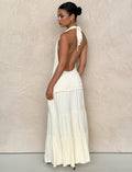 A woman poses sideways against a neutral textured wall wearing the TIGERMIST DENALI HALTER DRESS in Bone, a fitted cream-colored maxi with a tiered skirt. Her hair is styled neatly in a bun.