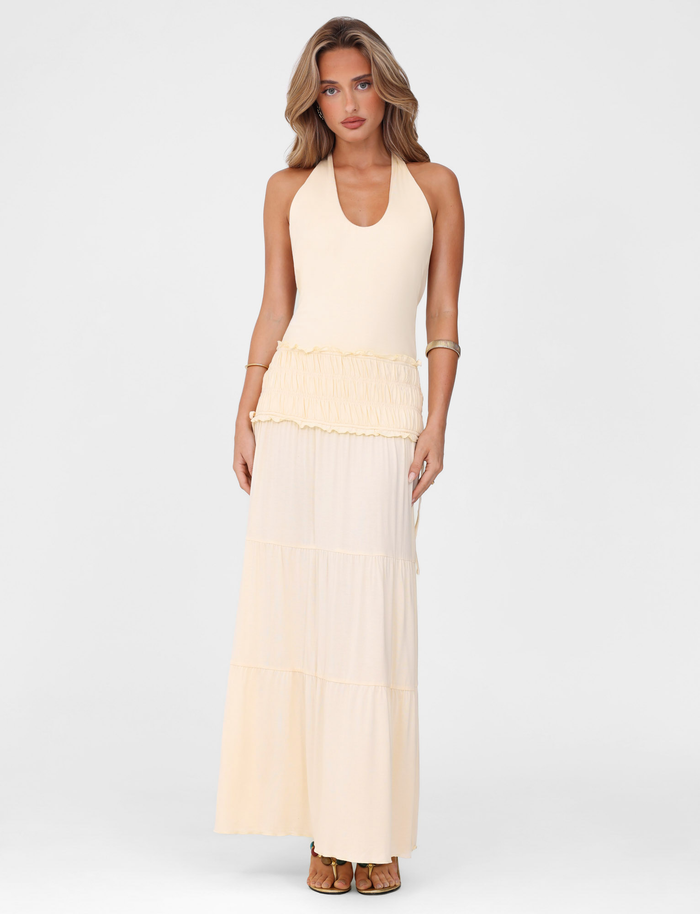 A woman with wavy light brown hair wears the TIGERMIST DENALI HALTER DRESS - NEUTRAL : BONE, a sleeveless, cream maxi with a tiered skirt and ruffled waist. She stands against a plain white background, accessorized with a gold bracelet and sandals.