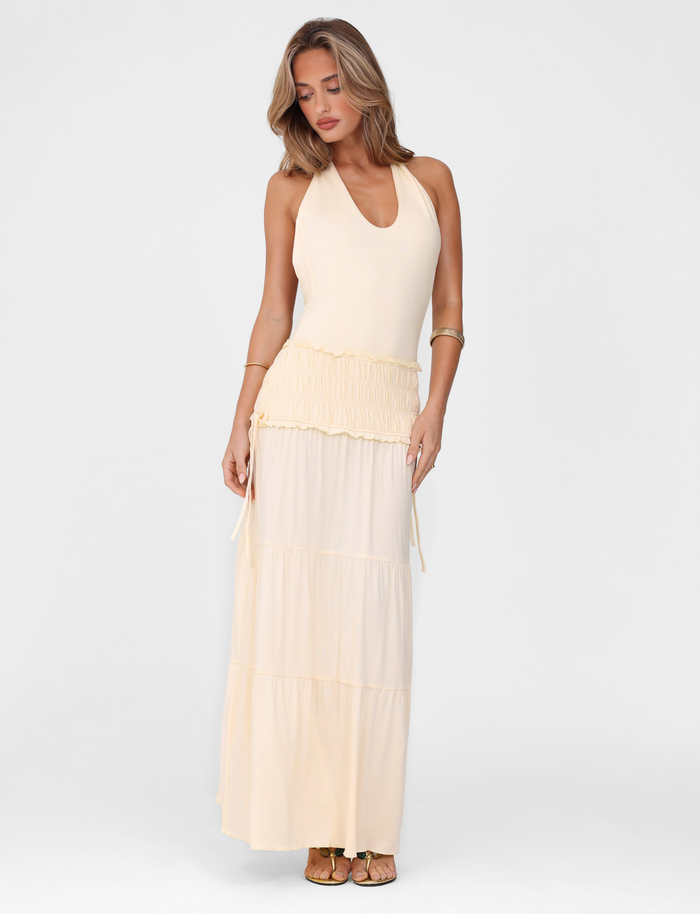 A woman wearing the TIGERMIST DENALI HALTER DRESS in Neutral : Bone stands against a plain white background, looking down with one hand by her side and the other holding her skirt, paired with gold sandals.