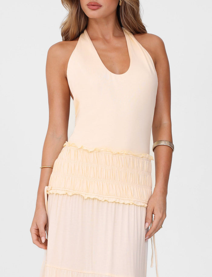 A woman wears the TIGERMIST DENALI HALTER DRESS in Bone, a sleeveless maxi dress with a gathered waistband and flowing skirt, styled with gold bangles and loose waves.