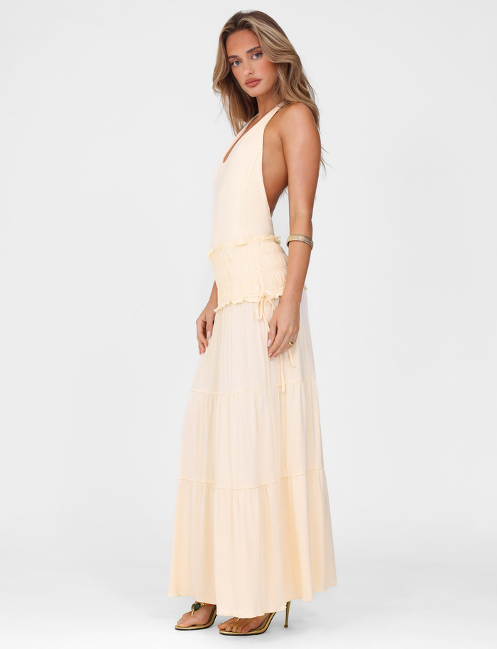 A woman with long, wavy hair models the TIGERMIST DENALI HALTER DRESS in Neutral: Bone—a fitted jersey maxi with a tiered skirt. She stands sideways in high-heeled sandals against a plain white background.