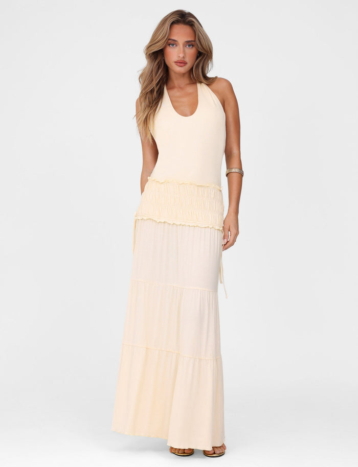 A woman with long, wavy hair wears the TIGERMIST DENALI HALTER DRESS in Neutral Bone—a fitted sleeveless maxi with a tiered skirt—paired with beige sandals, standing against a plain white background.